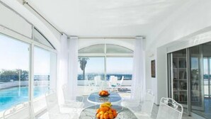 Ein modernes Zimmer mit einem Glastisch, weißen Stühlen und Blick auf Pool und Meer.