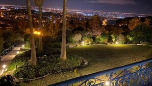Ein nächtlicher Blick auf eine Stadtlandschaft mit einem gut beleuchteten Gartenbereich, der einen Weg und Palmen zeigt.
