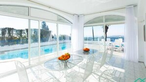 Ein modernes Zimmer mit einem Glastisch und Stühlen, Blick auf einen Swimmingpool und einem Balkon mit Liegestühlen.