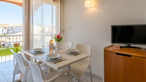 A dining area with a table set for two, a TV on a wooden cabinet, and a balcony with a view of buildings. - Primavera by Interhome (Cap d'Agde)