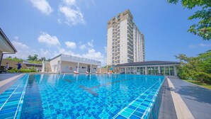 A swimming pool with clear blue tiles, surrounded by a building with a glass facade and a tall residential structure in the background. - Galle Holiday Home Rental - 3BR, Fast Internet, Pool, Gym, Tennis, Beach House  (Galle)
