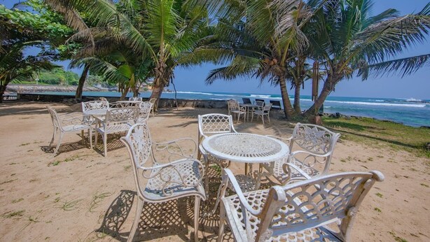 A beachside seating area with white metal chairs and tables, surrounded by palm trees and overlooking the ocean. - Galle Holiday Home Rental - 3BR, Fast Internet, Pool, Gym, Tennis, Beach House  (Galle)