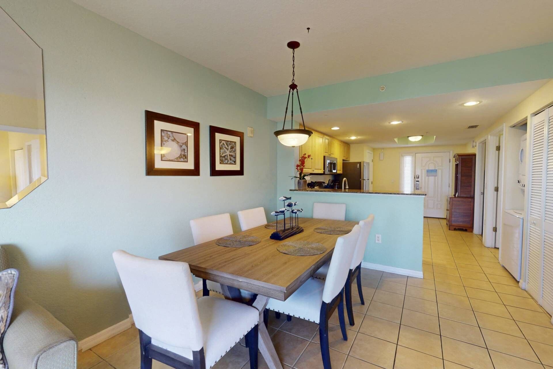 Un comedor con mesa de madera y sillas blancas, una cocina al fondo y un pasillo que conduce a una puerta.