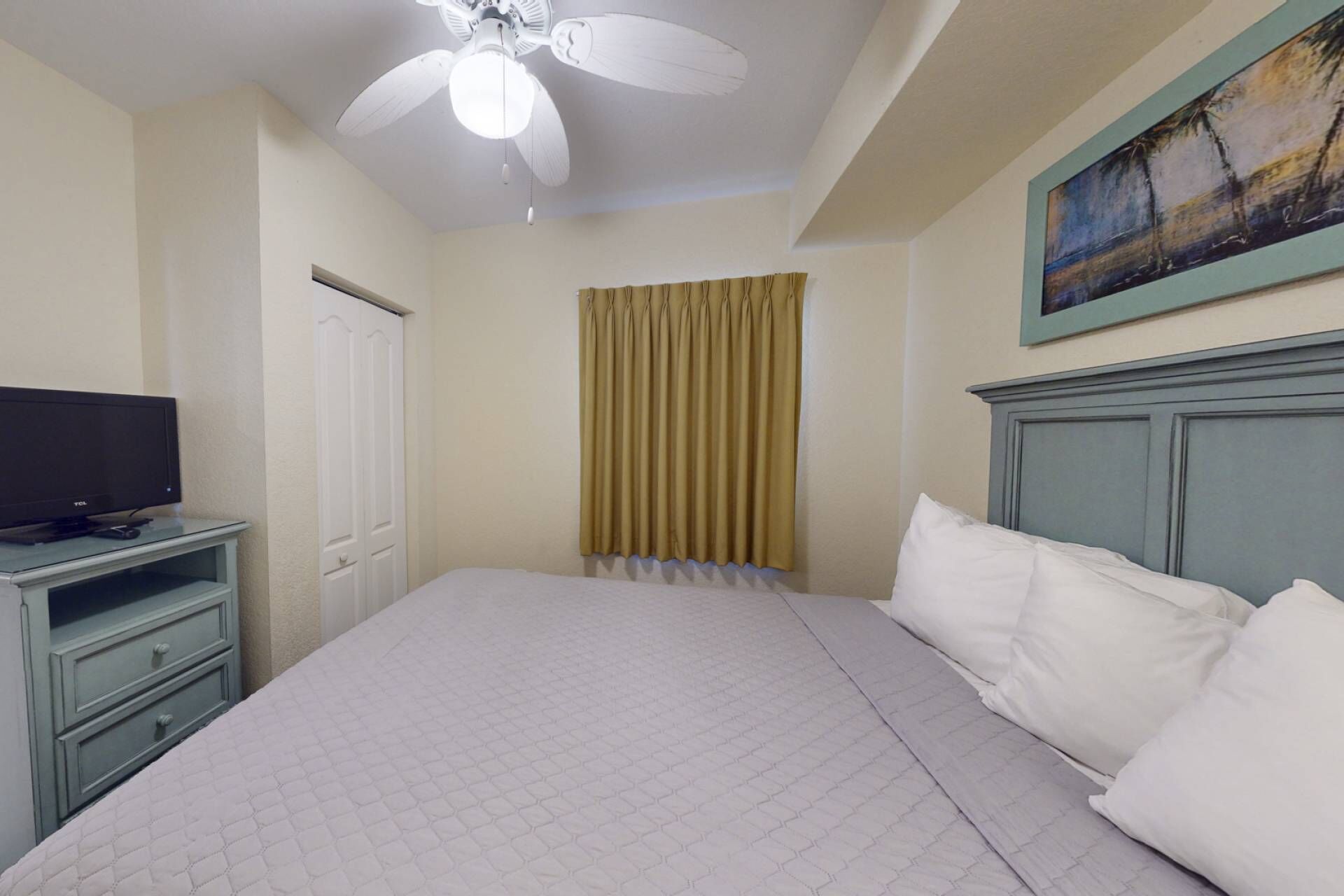 Habitación de hotel con cama, televisor sobre un soporte, ventilador de techo, puerta y un cuadro en la pared.