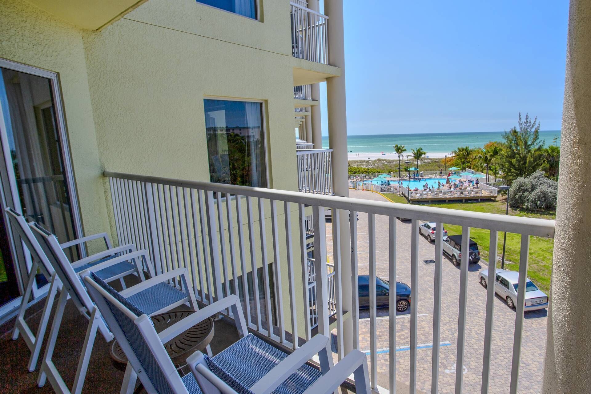 Un balcon avec des chaises blanches, donnant sur une piscine et une plage.