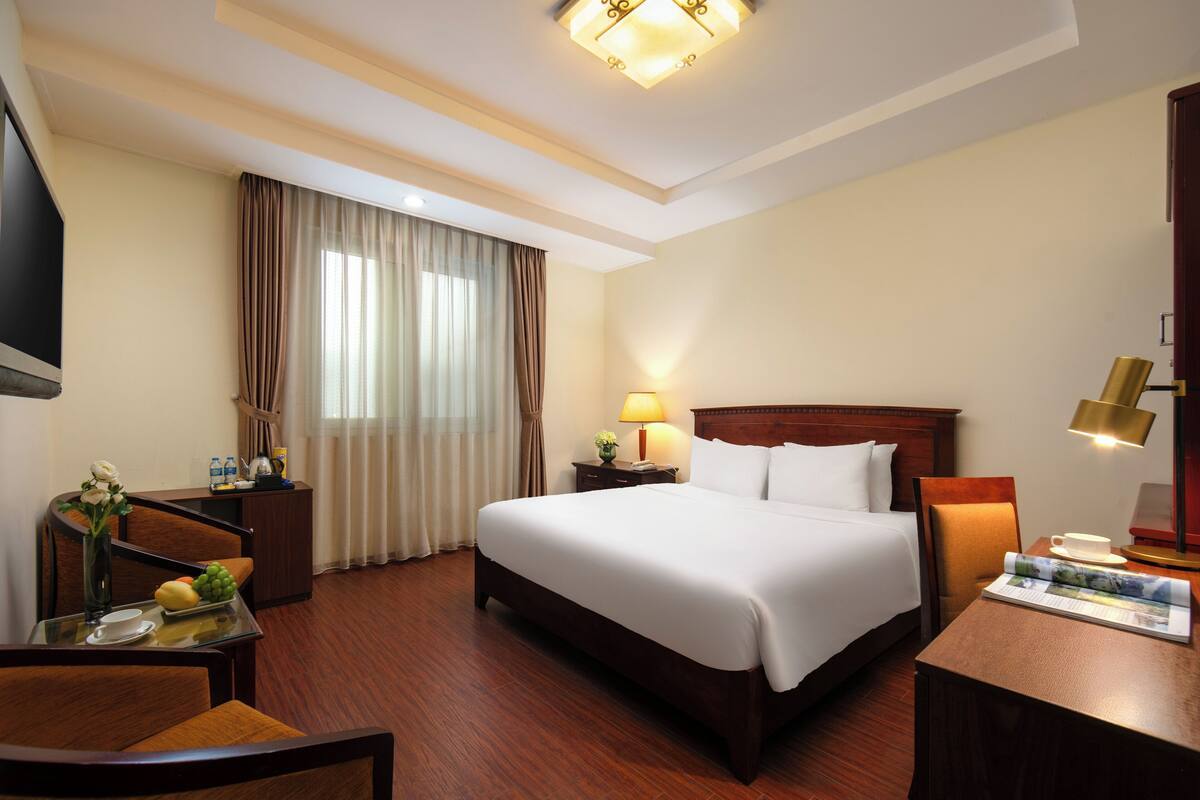 Ein Hotelzimmer mit einem großen Bett, einem Schreibtisch mit Lampe, einem Sessel, einem Fernseher und einem Fenster mit Vorhängen.