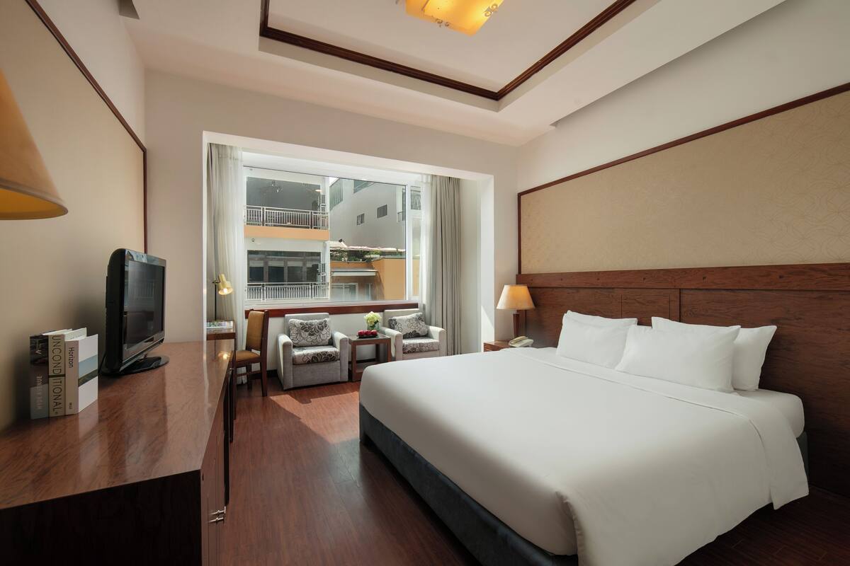 Ein Hotelzimmer mit einem großen Bett, einem Schreibtisch mit Fernseher, einer Sitzecke mit Stühlen und Blick auf die Außenansicht des Gebäudes durch ein großes Fenster.