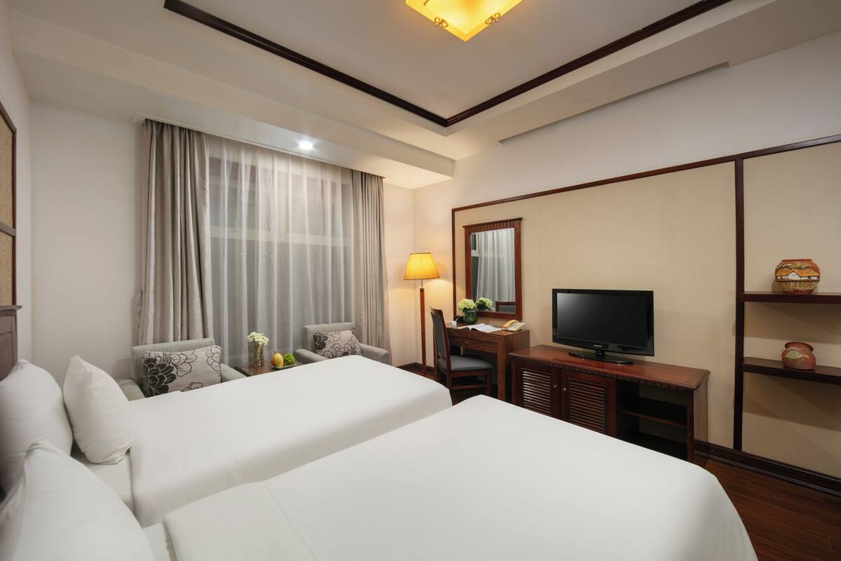 Ein Hotelzimmer mit zwei Betten, einem Schreibtisch, einem Fernseher, einer Lampe und einem großen Fenster mit Vorhängen.