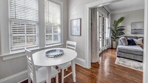 A dining area with a white table and chairs, large windows with blinds, a grey sofa, and a potted plant.