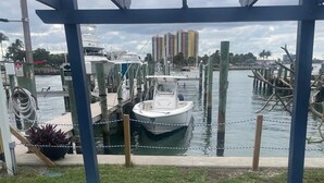 Un área de estar al aire libre techada con sillas Adirondack y una mesa, con vista a un puerto deportivo con barcos y edificios al fondo.