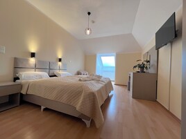 Quarto com cama, mesas de cabeceira, escrivaninha, televisão e janela.
