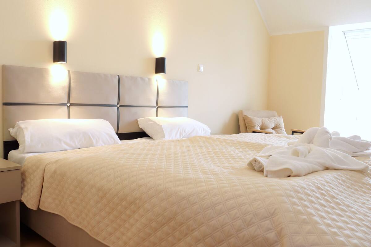 A neatly made bed with a quilted bedspread, two bedside tables, a chair, and wall-mounted lighting.