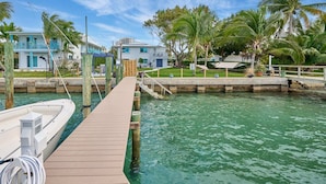 A wooden dock leading to a marina with clear turquoise water, surrounded by palm trees and two buildings with blue balconies. - Elegant 2BR Haven by the Water (Riviera Beach)