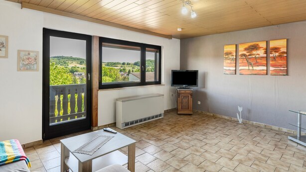 A room with a wooden ceiling, a large window with a view, a radiator, a TV, and a painting on the wall.
