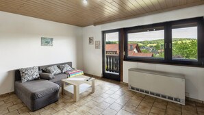 A room with a grey sofa, a small table, a radiator, and a large window with a view of buildings and greenery.