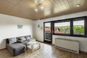 Une pièce avec un canapé gris, une petite table, un radiateur et une grande fenêtre donnant sur des immeubles et de la verdure.