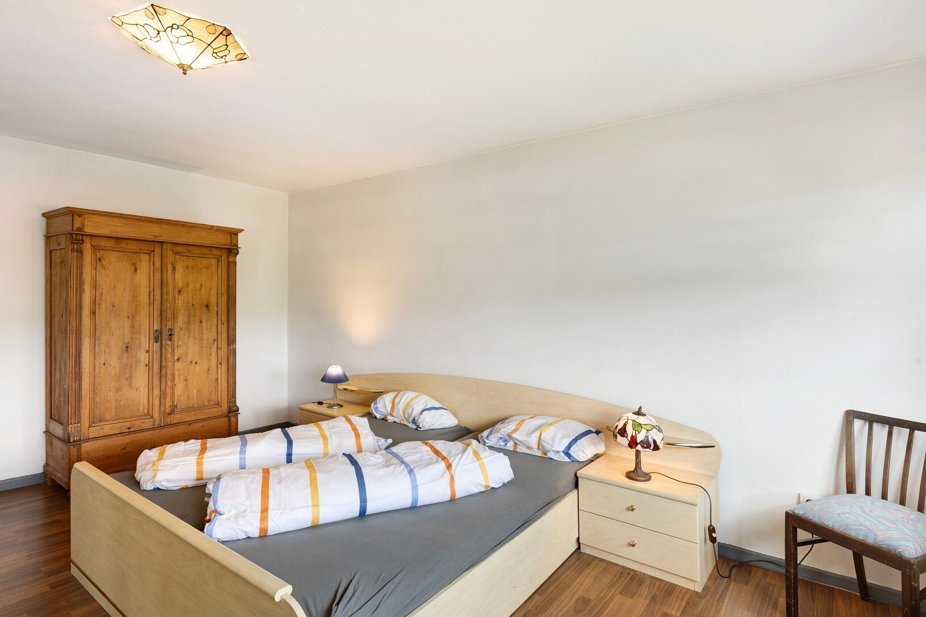 Un dormitorio con cama, armario de madera, mesita de noche, lámpara, silla y aplique de pared.