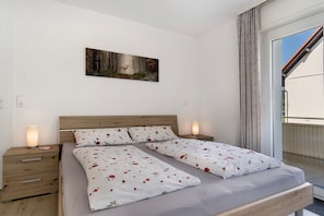 Una cama doble con ropa de cama floral, una mesita de noche con lámpara, un cuadro de un paisaje boscoso y vistas de un edificio a través de una ventana.