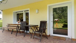 Un patio exterior con mesa y sillas, un banco, una puerta con vistas a follaje y un comedero para pájaros sobre un trípode.