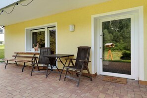 An outdoor patio with a table and chairs, a bench, a door with a view of greenery, and a birdhouse on a tripod.