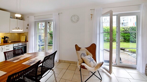 Een keuken met kasten, een eettafel en stoelen. Er hangt een klok aan de muur en een stoel met een stervoorkussen.