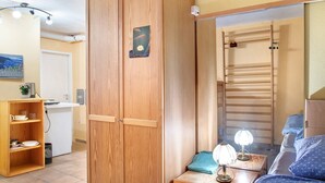 A room with a wooden wardrobe, a small desk with a lamp, a bed with blue bedding, and a painting on the wall.