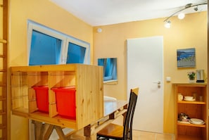 Una habitación con literas, una mesa de comedor y una silla. Hay una puerta, un estante con libros y una planta, y una ventana con cortinas.