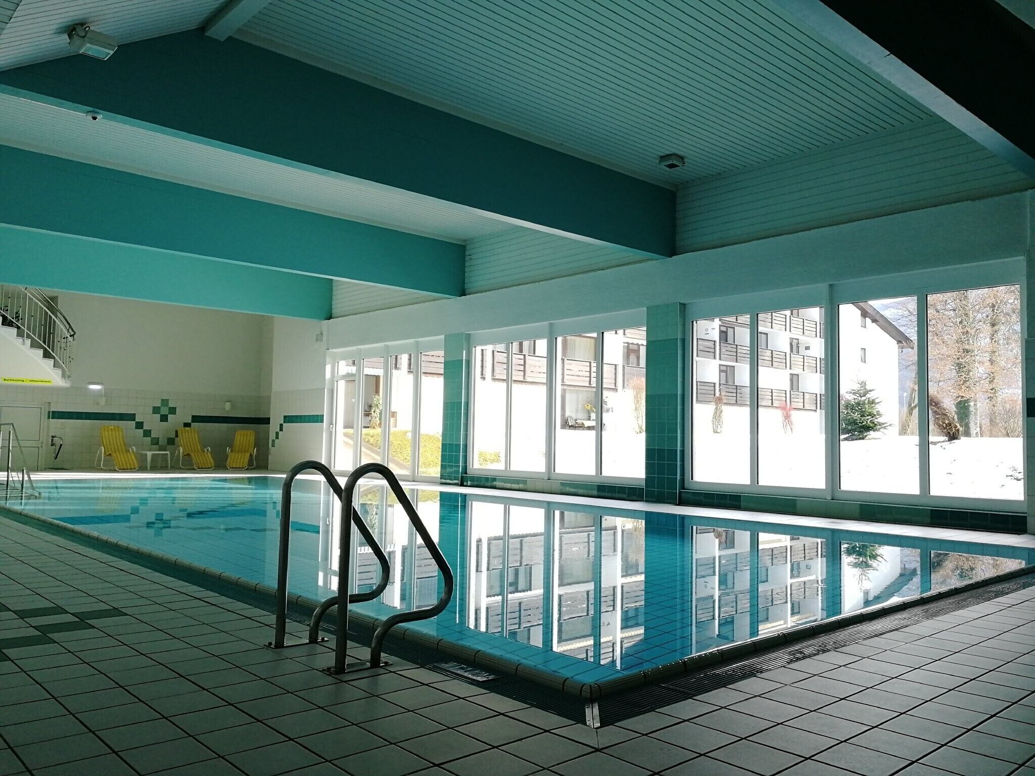 Une piscine intérieure avec des parois en verre transparent, un sol carrelé et un escalier menant à un niveau supérieur.