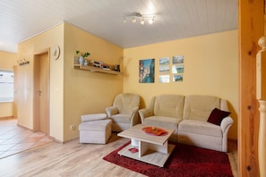 Uma sala de estar com sofá, duas poltronas, mesa de centro, tapete vermelho, prateleira com livros e um vaso, e uma porta.