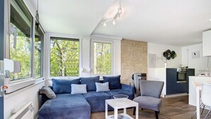 Una sala de estar moderna con un sofá seccional, una mesita blanca y un sillón gris. Se observa una zona de cocina con estufa y alacenas.