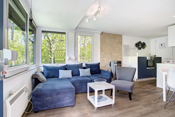 A modern living room with a sectional sofa, a small white table, and a grey armchair. There is a kitchen area with a stove and cabinets.