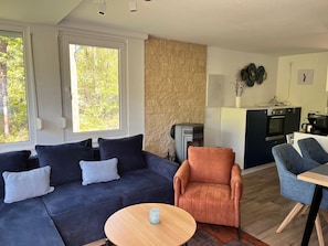 A modern living room with a sofa, armchair, and coffee table. A stone wall and kitchen with appliances are visible.