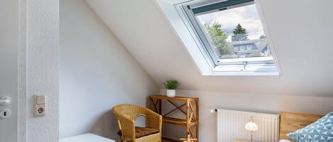 Un dormitorio con techo inclinado, tragaluz, cama con ropa de cama estampada, mesita de noche con lámpara, una silla de mimbre y una pequeña mesa blanca.