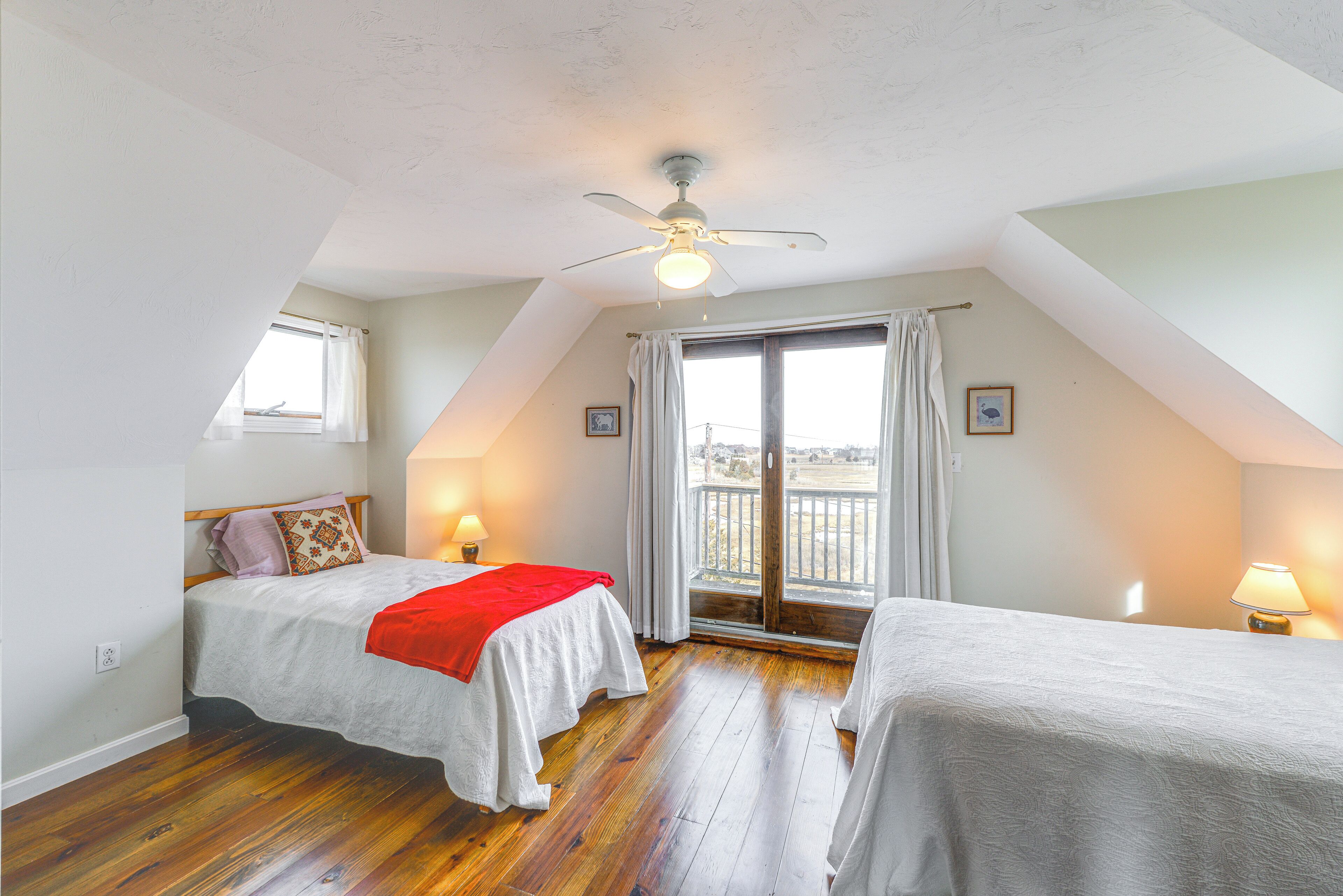 Ein Schlafzimmer mit zwei Betten, einem Deckenventilator, einem Fenster mit Vorhängen und Balkonblick.