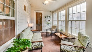 A sunlit room with a wooden floor, white walls, and large windows. There are two metal-framed chairs, a sofa, and a small table with a plant. - Historical 1835 Home in Grand Coteau! (Grand Coteau)