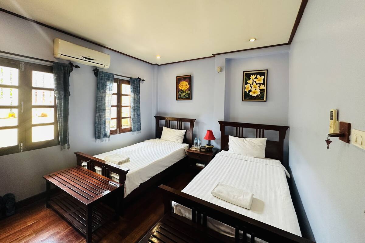 A room with two beds, a bench, an air conditioning unit, a window with curtains, and a wall-mounted light switch.