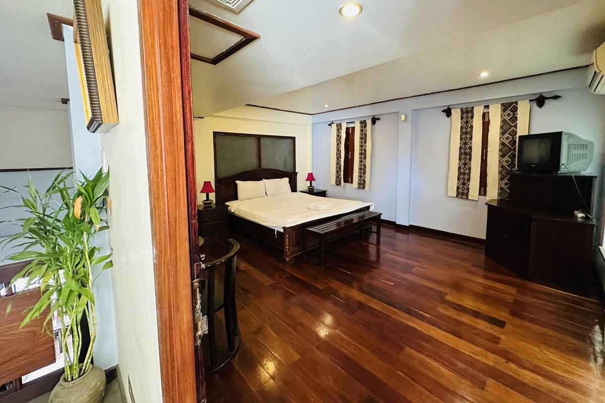 A spacious bedroom with a large bed, wooden flooring, a potted plant, a television, and patterned curtains.