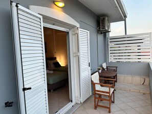 Een balkon met een tafel en stoelen, een open deur die leidt naar een slaapkamer met een bed en kussens, en een airconditioner aan de muur bevestigd.