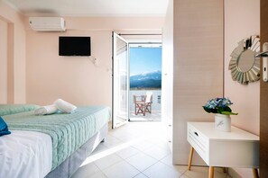 Quarto com cama, televisão, porta de correr para uma varanda e uma pequena mesa com um vaso.