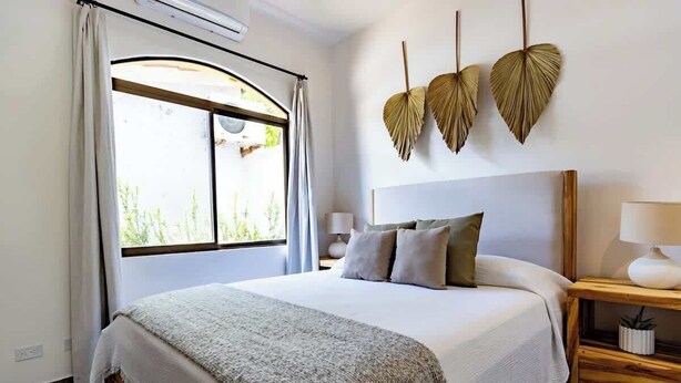 Een slaapkamer met een groot bed, een raam met gordijnen, een nachtkastje met een lamp en decoratieve wandkunst.
