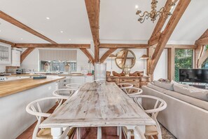 A spacious kitchen with wooden beams, a dining area with a long table and white chairs, and a cozy seating area with a sofa and TV. - Hiron's Piece and Honeysuckle Cottage with Hot Tub (Weston Subedge)