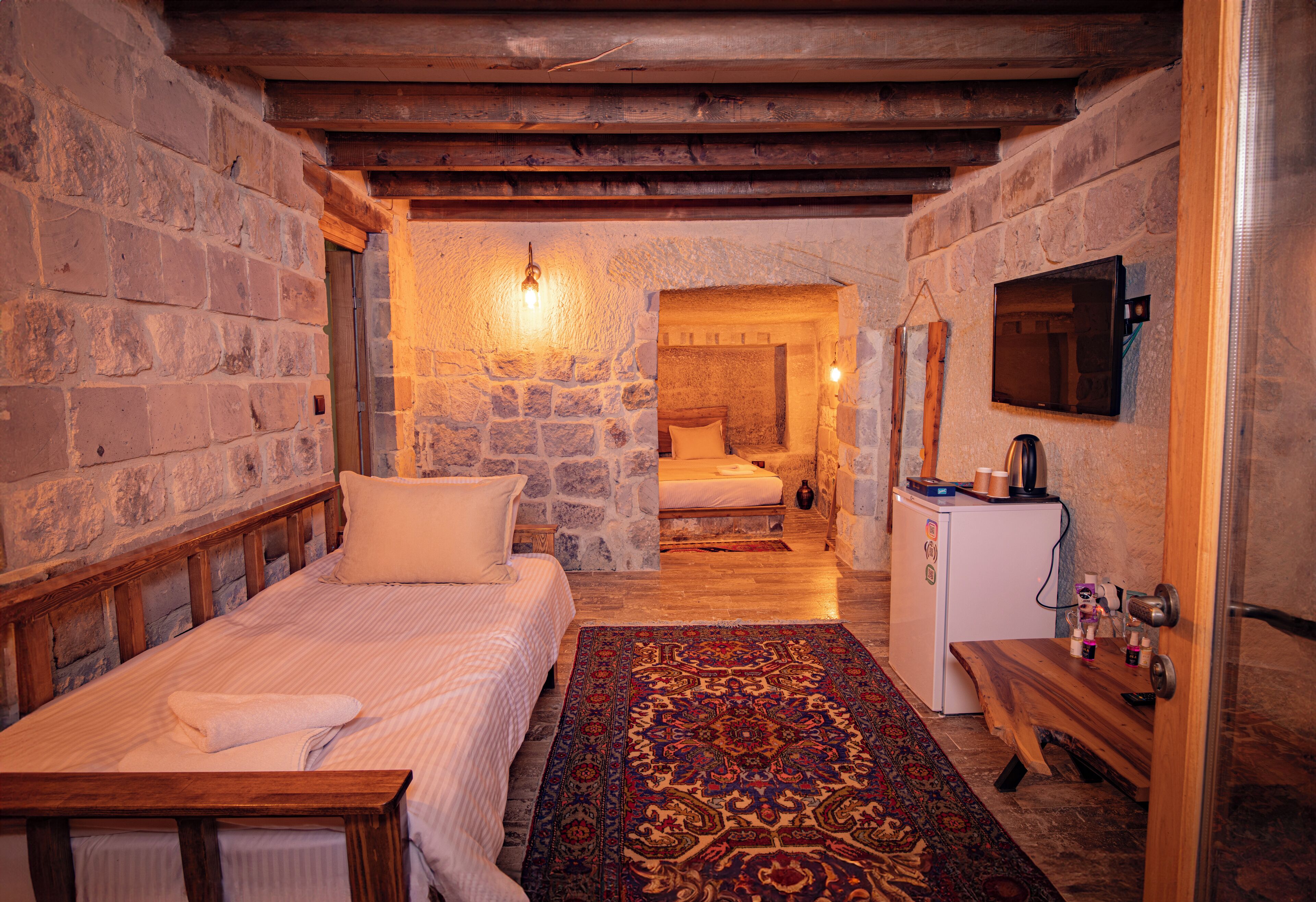 A cozy room with stone walls, a wooden bed, a small desk, and a television on the wall.