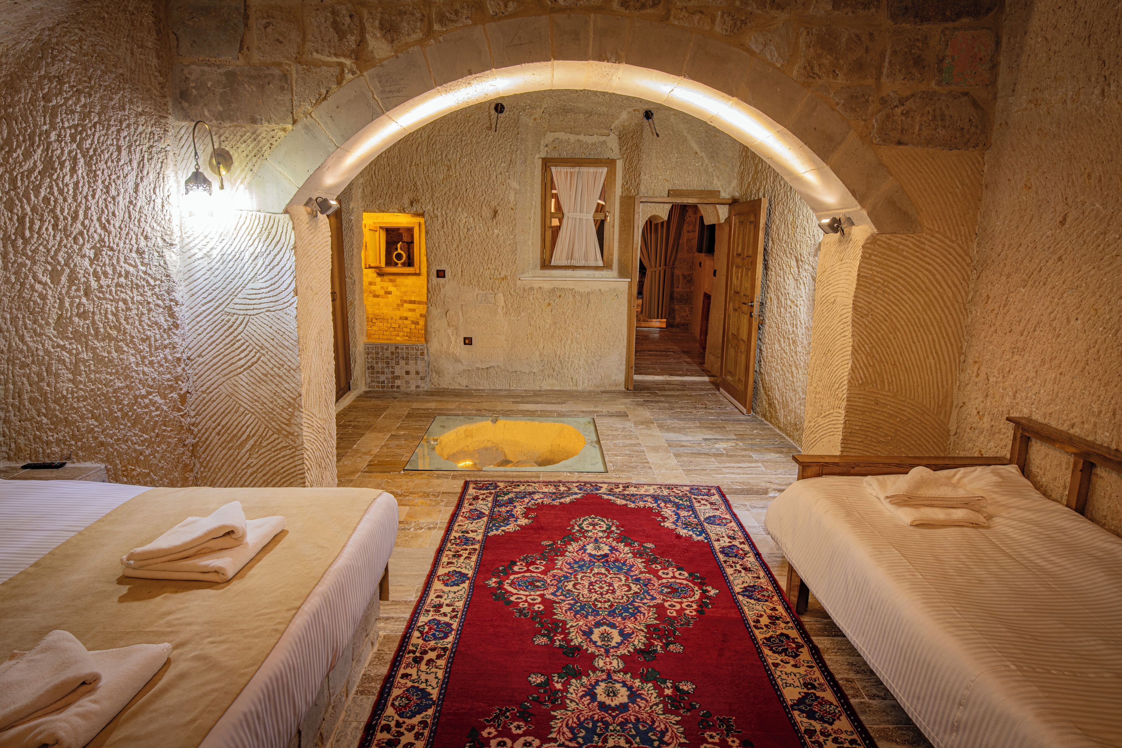 A room with two beds, a red patterned rug, stone walls, and a small window with curtains.