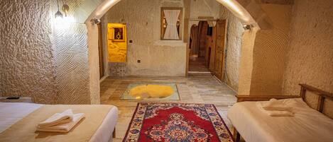 A room with two beds, a red patterned rug, stone walls, and a small window with curtains.