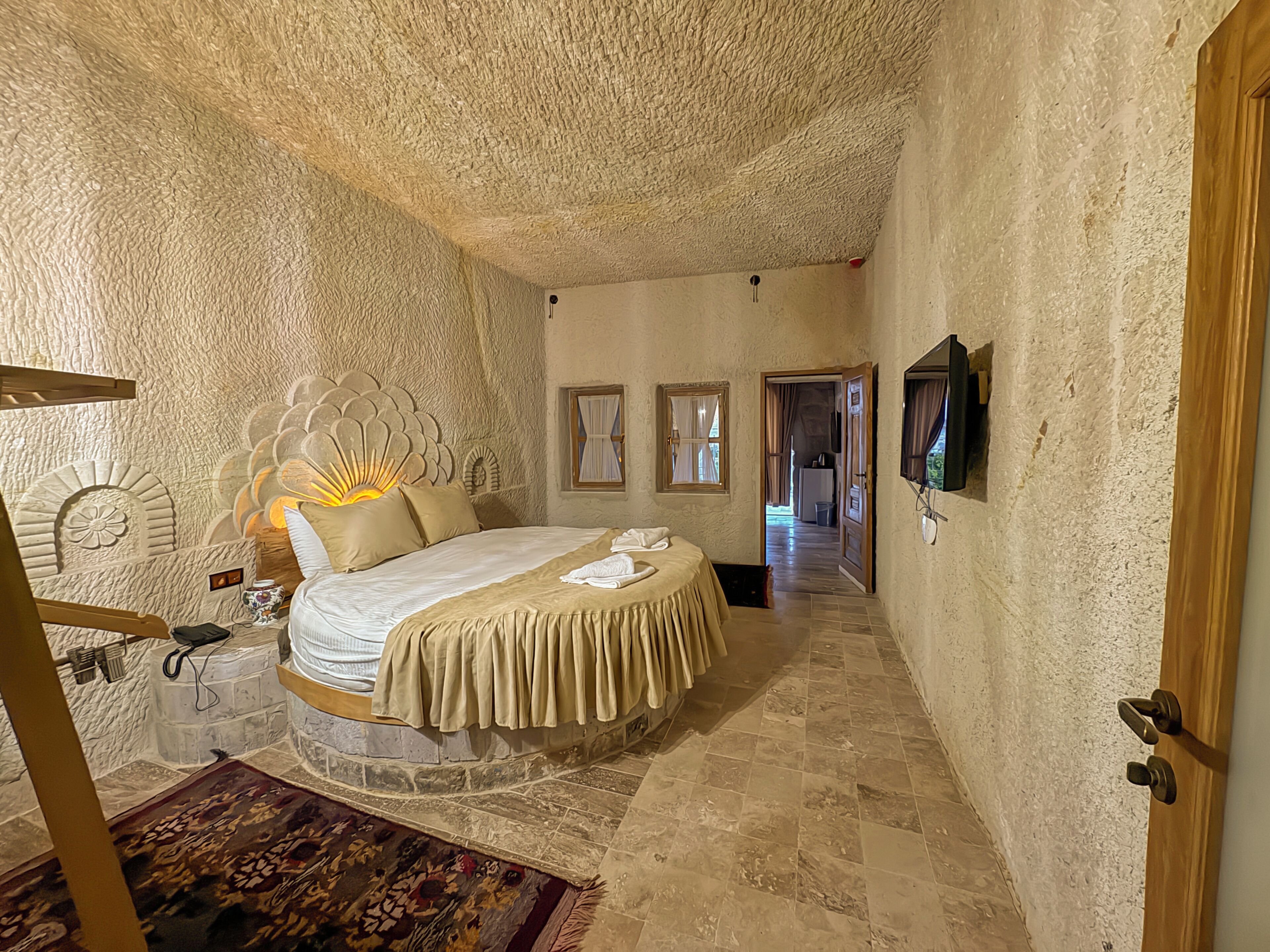 Un dormitorio con cabecera de piedra, una cama con colcha color beige, una puerta de madera y un tapete estampado.