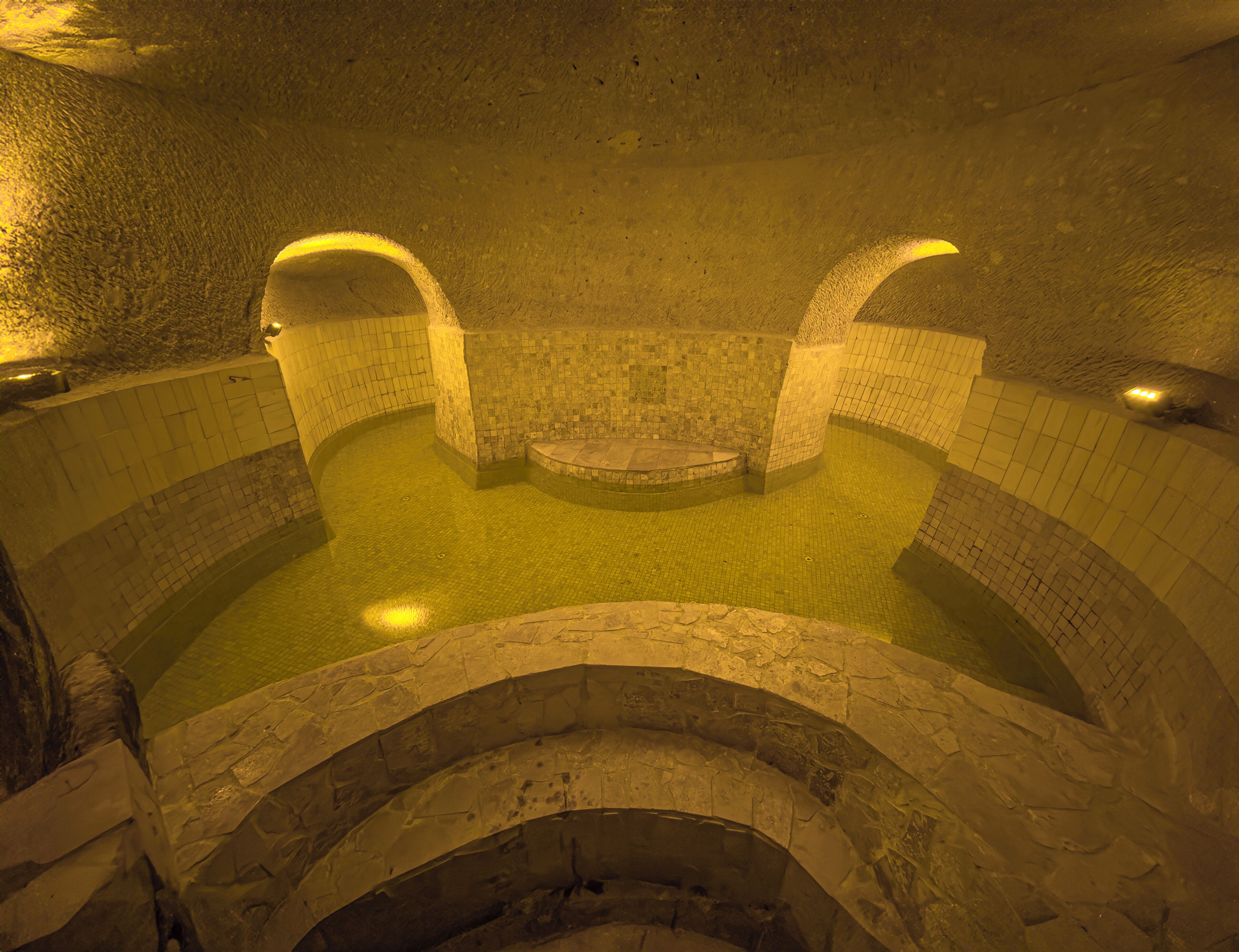 Un extenso baño termal circular con paredes de piedra y suelo de baldosas, iluminado con luz cálida.