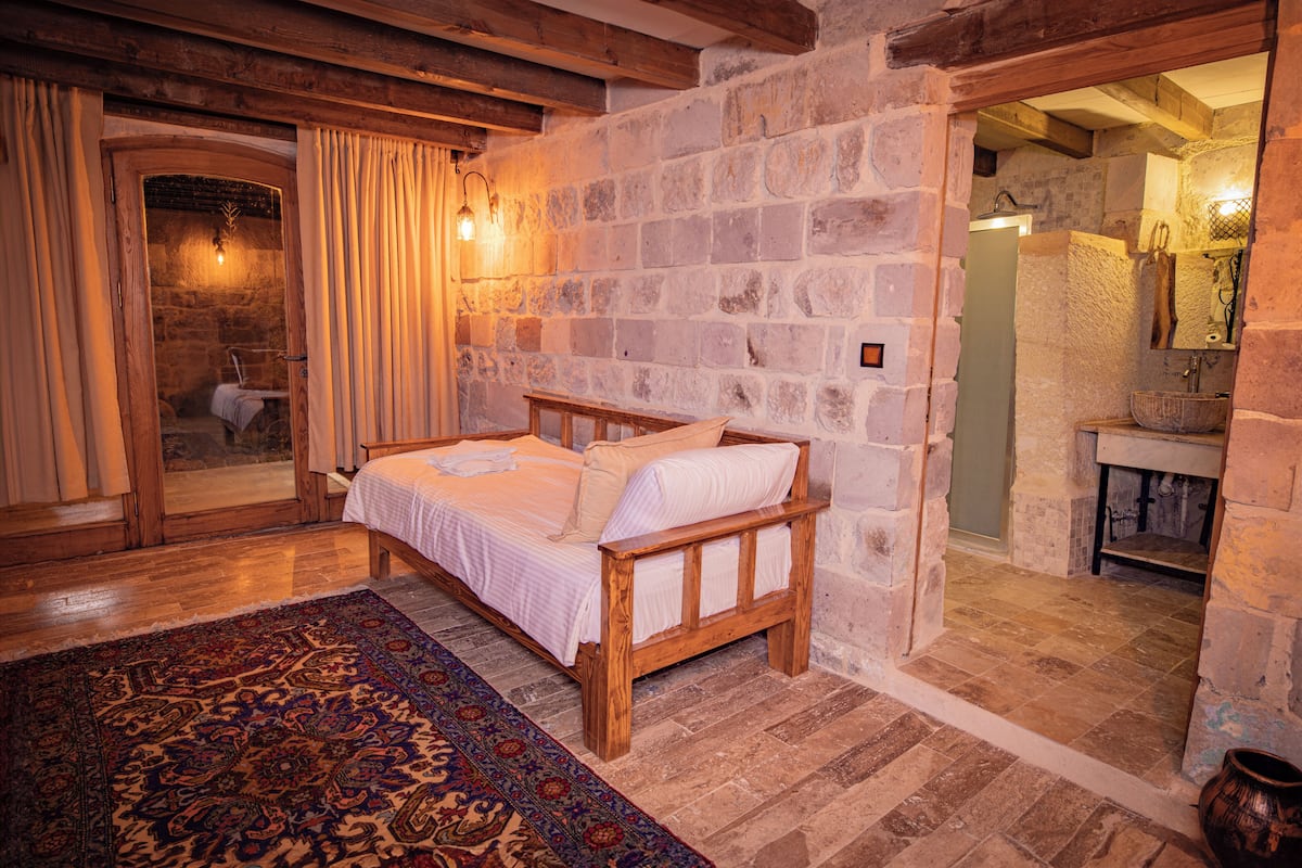Un lit en bois dans une pièce aux murs de pierre, agrémentée d’un tapis à motifs, d’un banc et d’une petite table.