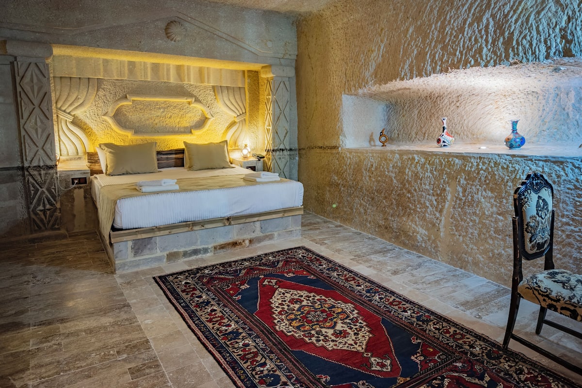 Une chambre avec un mur en pierre, un lit recouvert de draps blancs, un tapis à motifs et une table de chevet avec une lampe.