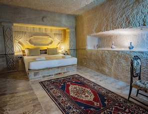A bedroom with a stone wall, a bed with white linens, a patterned rug, and a nightstand with a lamp. - Sur Cave Hotel (Ürgüp)