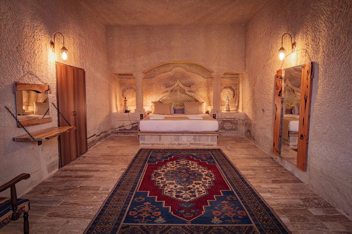 Une chambre à coucher avec un grand lit, un banc en bois, un tapis à motifs et des lampes fixées au mur.